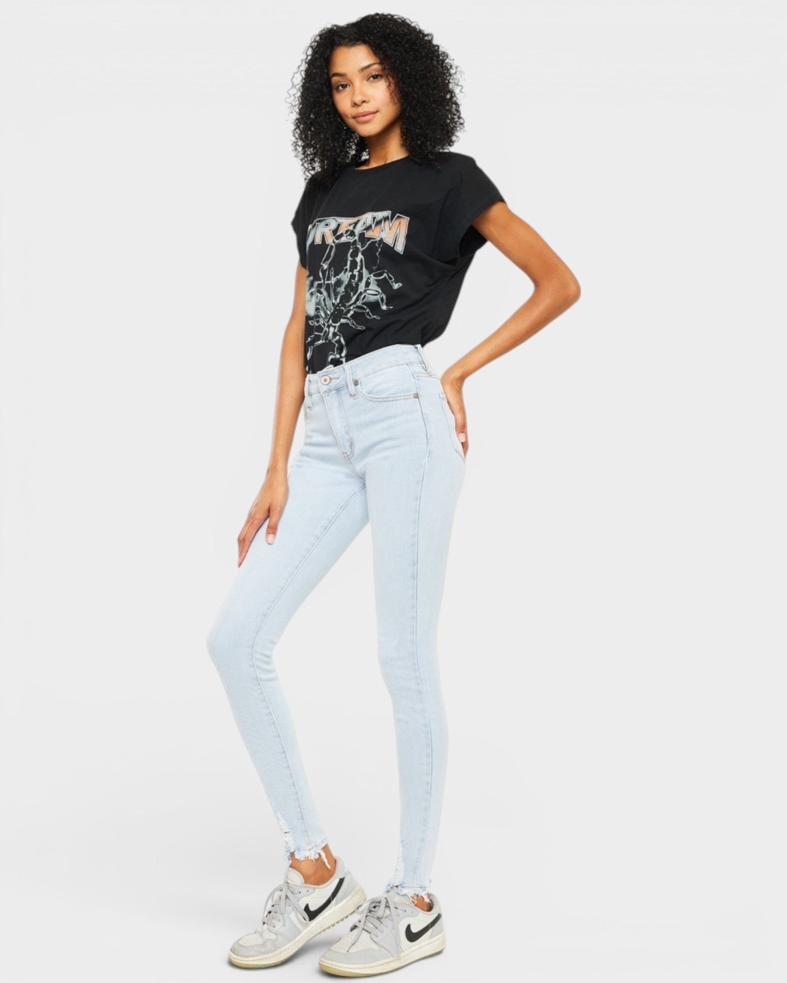 Woman wearing a black t-shirt with graphic design, light blue jeans, and white sneakers on a plain background