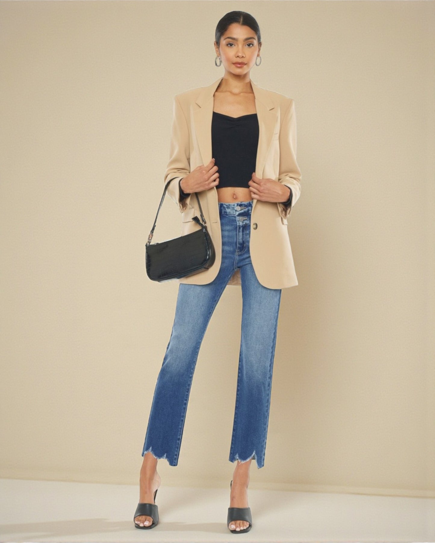 Woman wearing a beige blazer, black top, and blue jeans with a black handbag against a beige background
