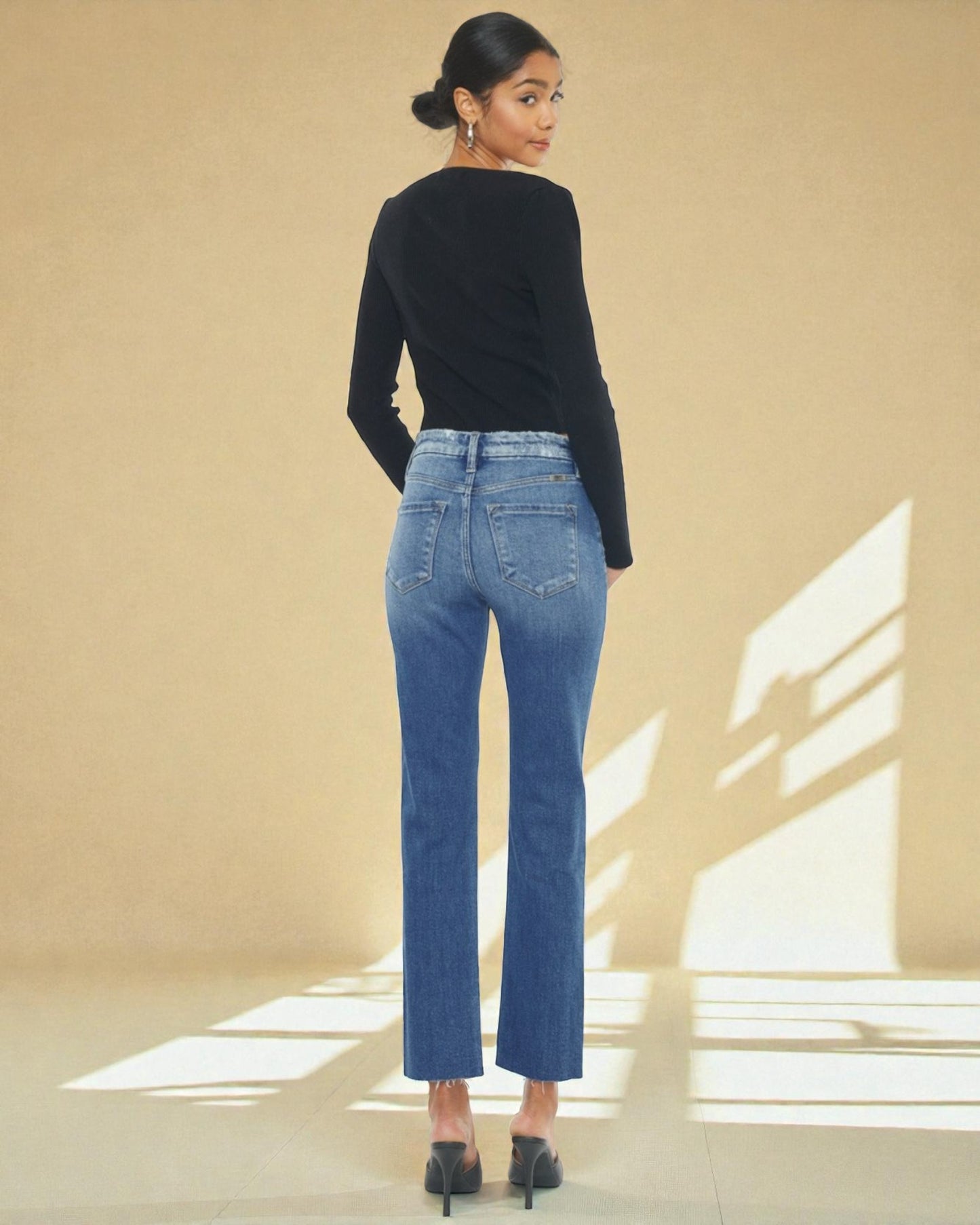 Woman wearing blue jeans and a black top standing in a sunlit room with a beige wall.