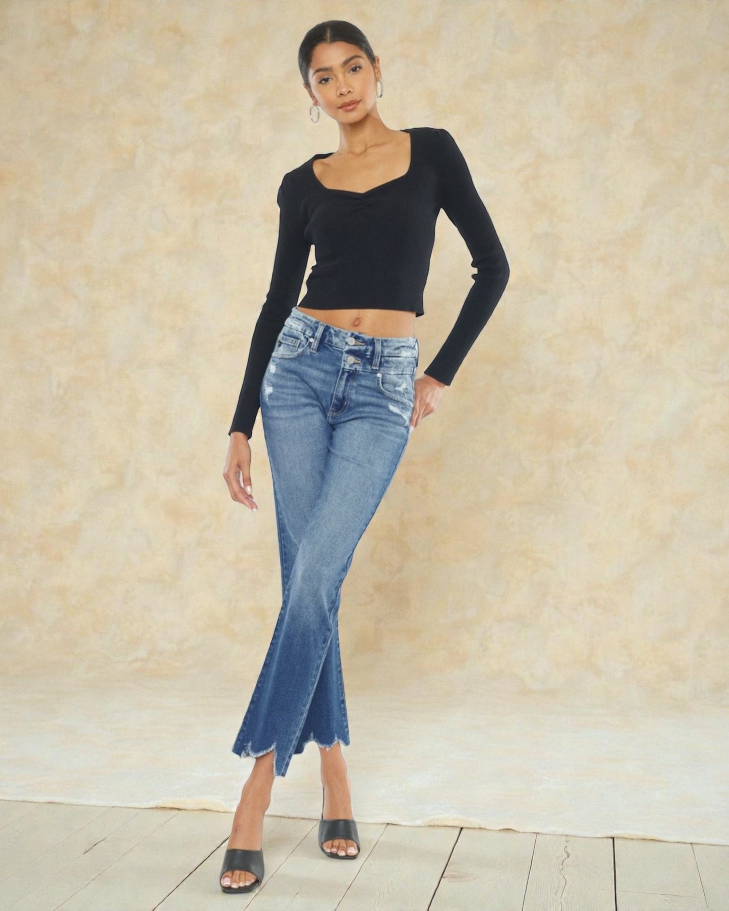 Woman wearing a black long-sleeve top and blue jeans against a beige wall.