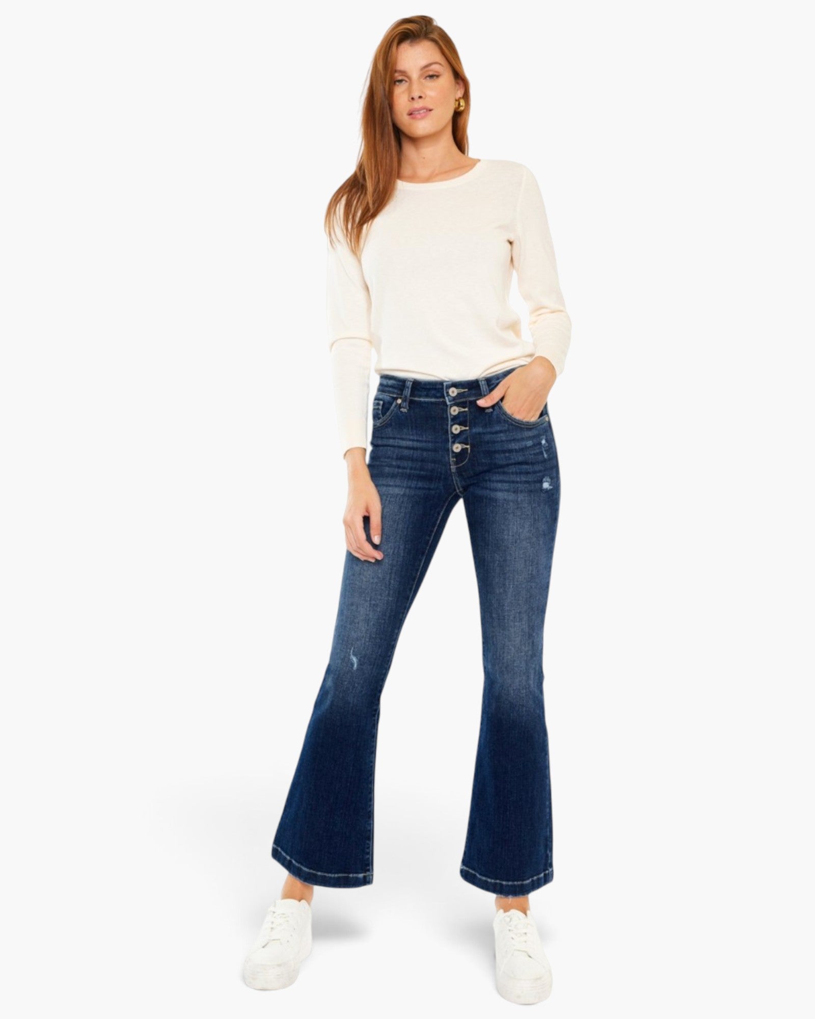 A woman modeling dark blue high-waisted flare jeans with a button fly, paired with a cream long-sleeve top and white sneakers, against a plain white background.