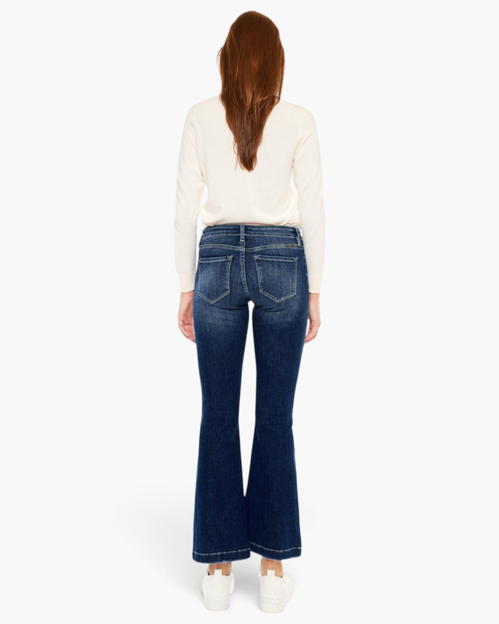 A woman viewed from the back wearing dark blue flared jeans, a cream long-sleeve top, and white sneakers against a plain white studio background.