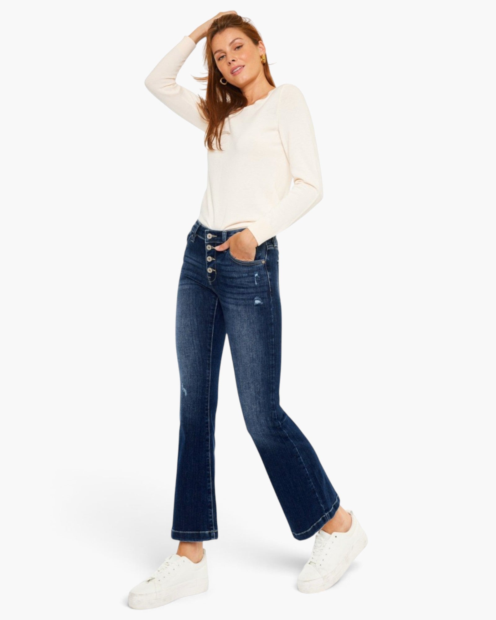 A woman modeling dark blue flare jeans with a white long-sleeve top and white sneakers, posing against a solid white studio background.