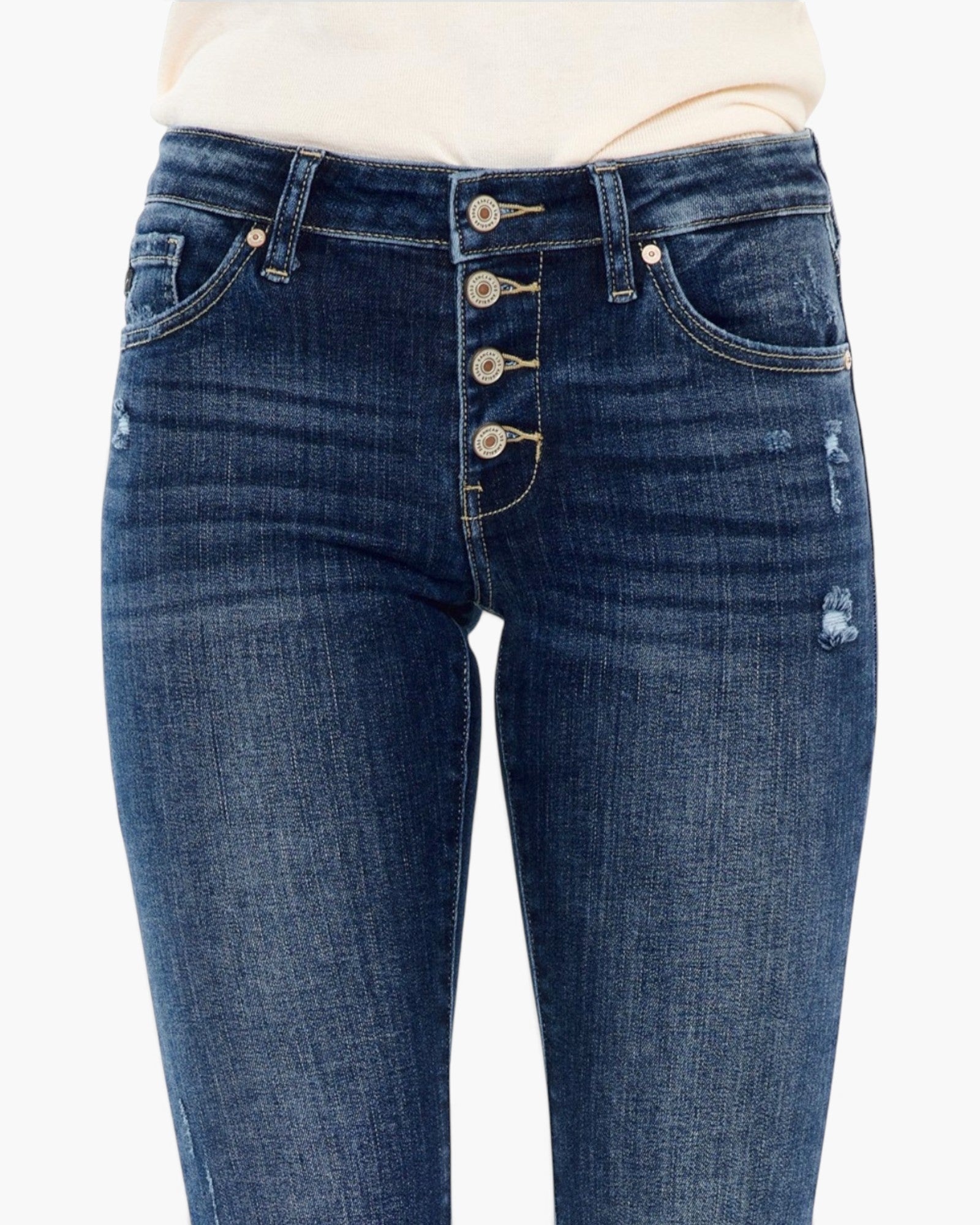 Close-up of a person wearing dark blue distressed denim jeans with a high waist and five front buttons, paired with a light cream top against a plain white background.