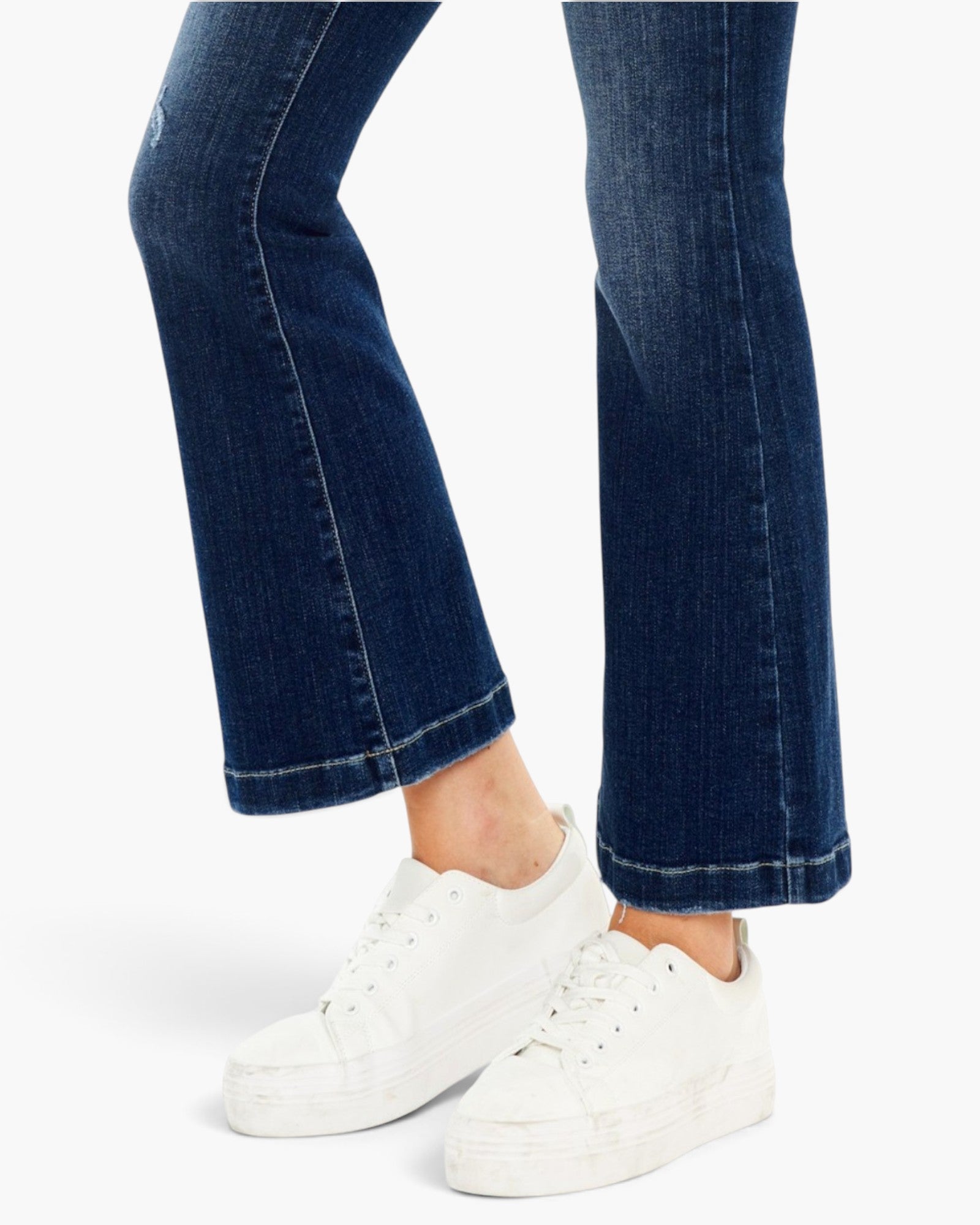 A close-up of a person's legs wearing dark blue flared denim jeans and white platform sneakers against a clean white background.