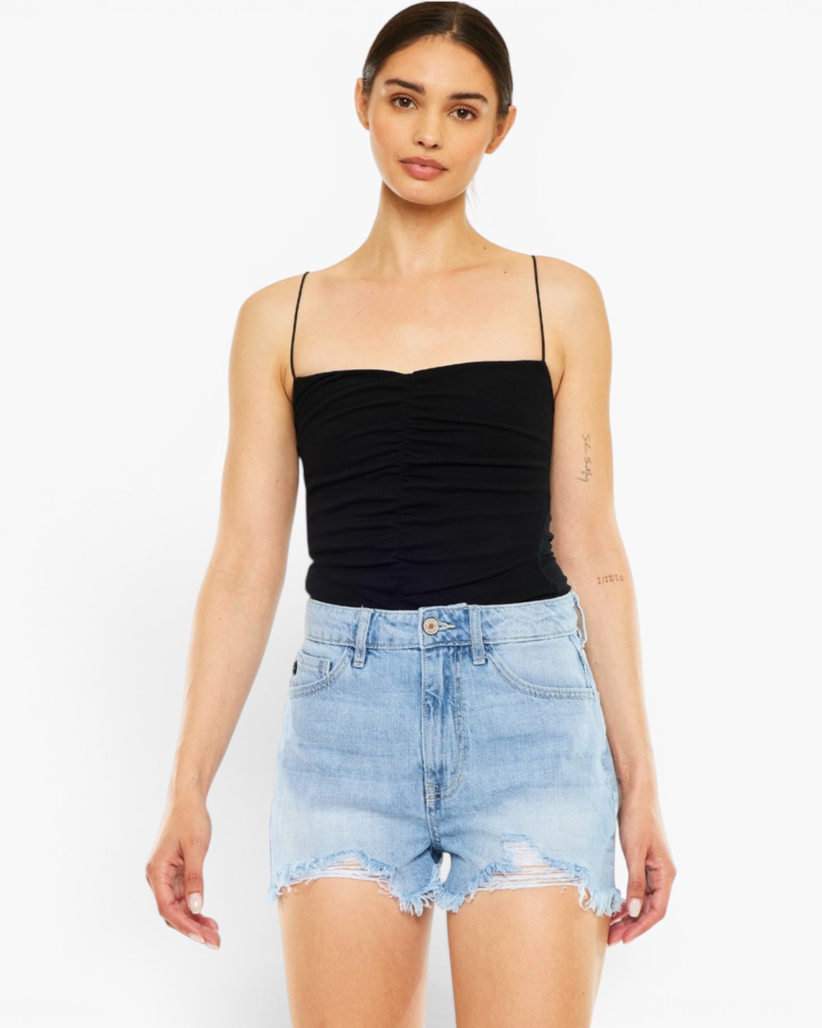 A woman modeling a black ruched spaghetti-strap camisole top paired with light-wash distressed denim shorts against a plain white studio background.