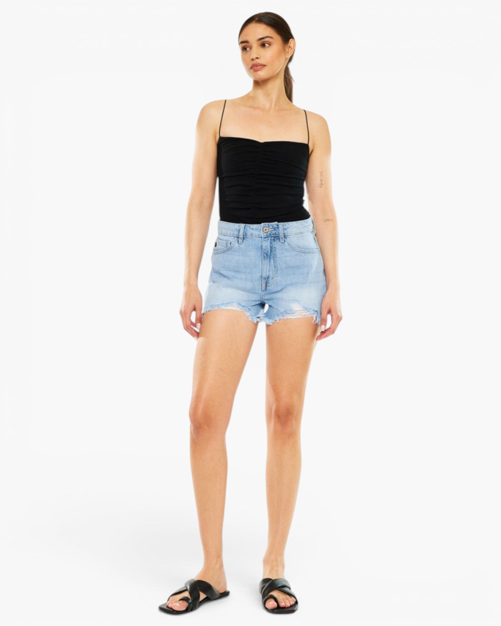 A woman modeling a black ribbed camisole top and light-wash denim shorts against a plain white studio background.