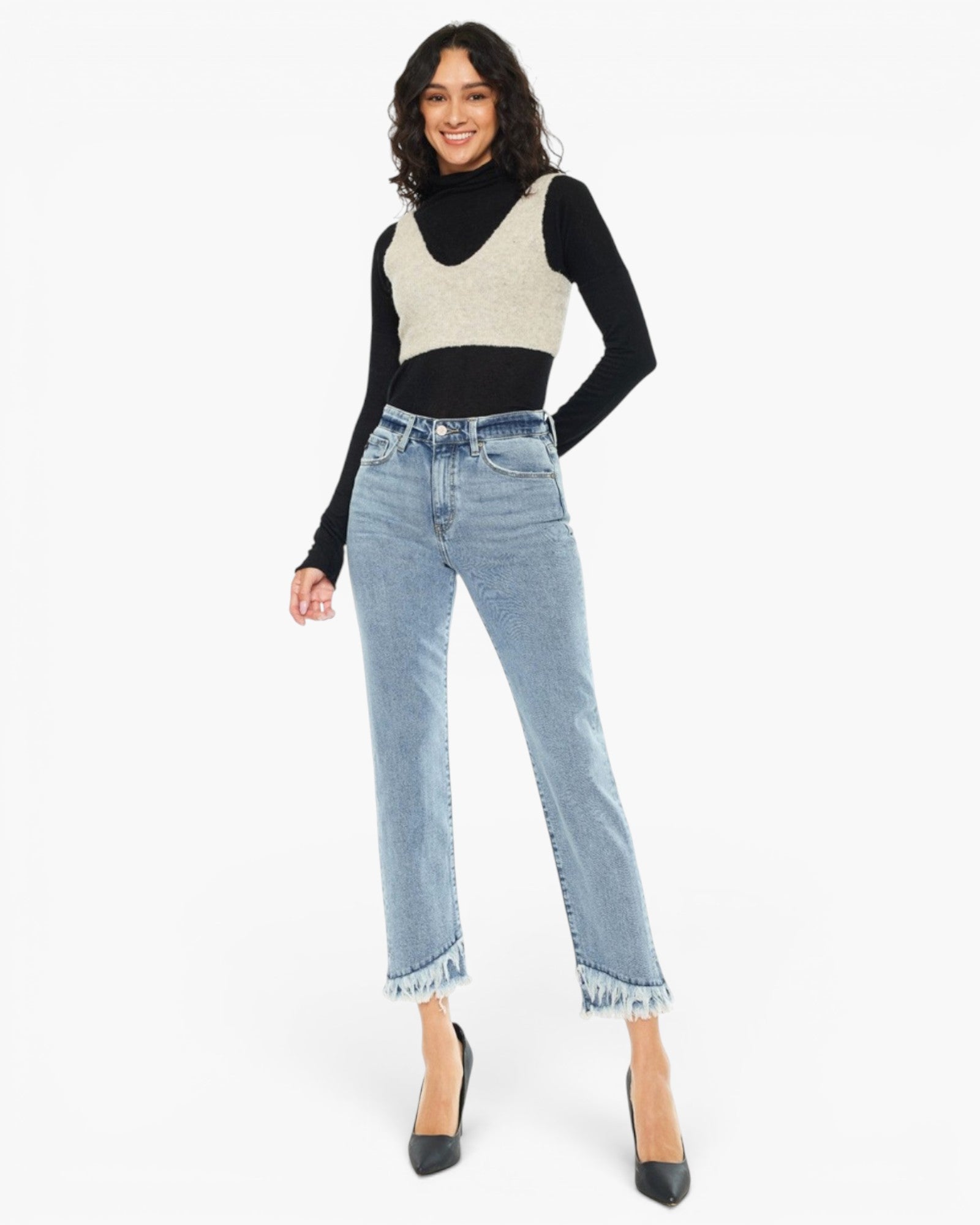 A model wearing a black long-sleeve turtleneck sweater layered under a cream knit vest, paired with light-wash frayed hem jeans and black pointed-toe heels, against a plain white studio background.