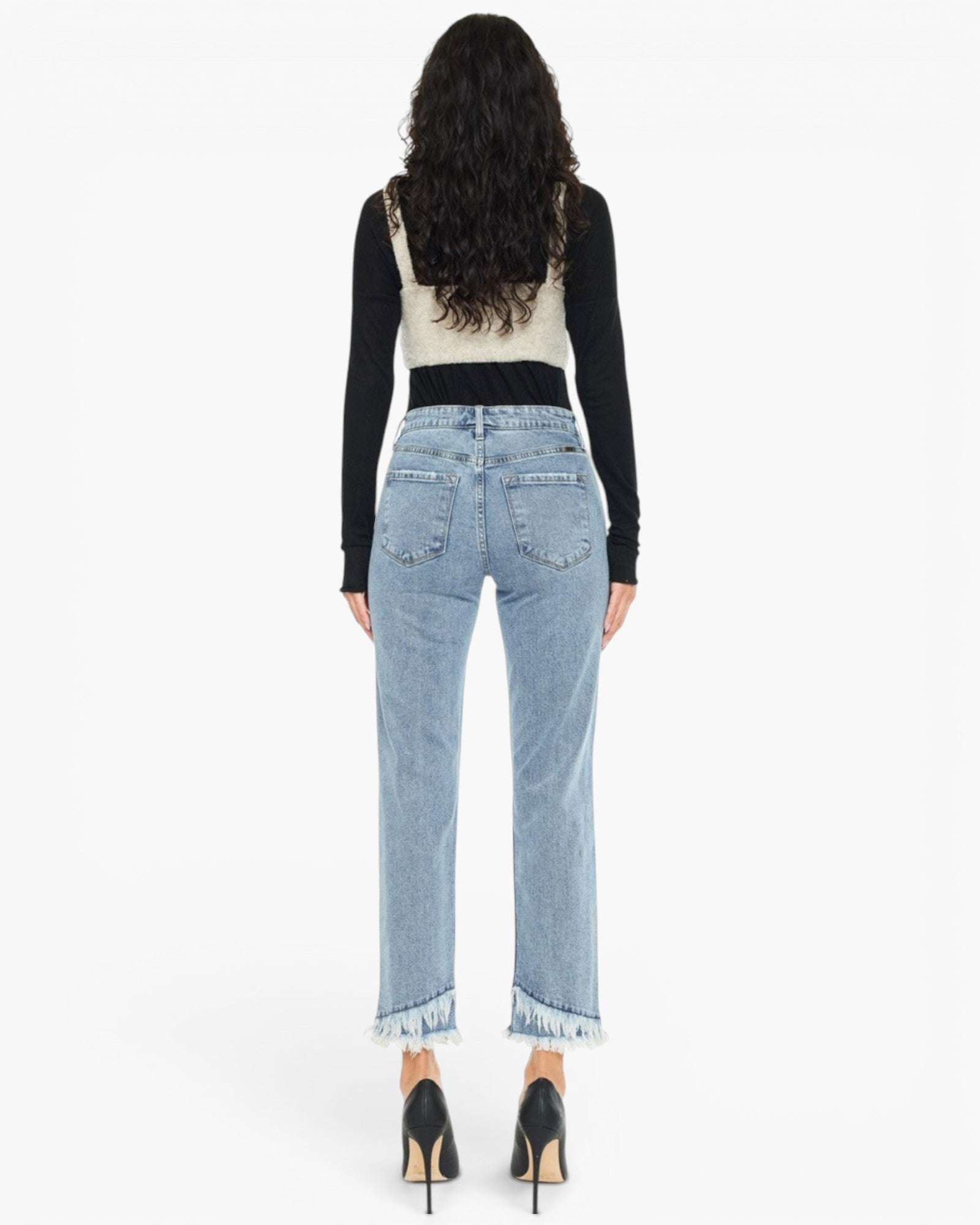 A woman shown from the back wearing light-wash blue cropped jeans with frayed hems, a black long-sleeve top layered over a cream vest, and black high heels against a plain white studio background.