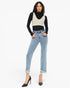 A model wearing a black long-sleeve turtleneck, a beige knit vest, light-wash blue cropped jeans with frayed hems, and black high heels, standing against a plain white studio background.