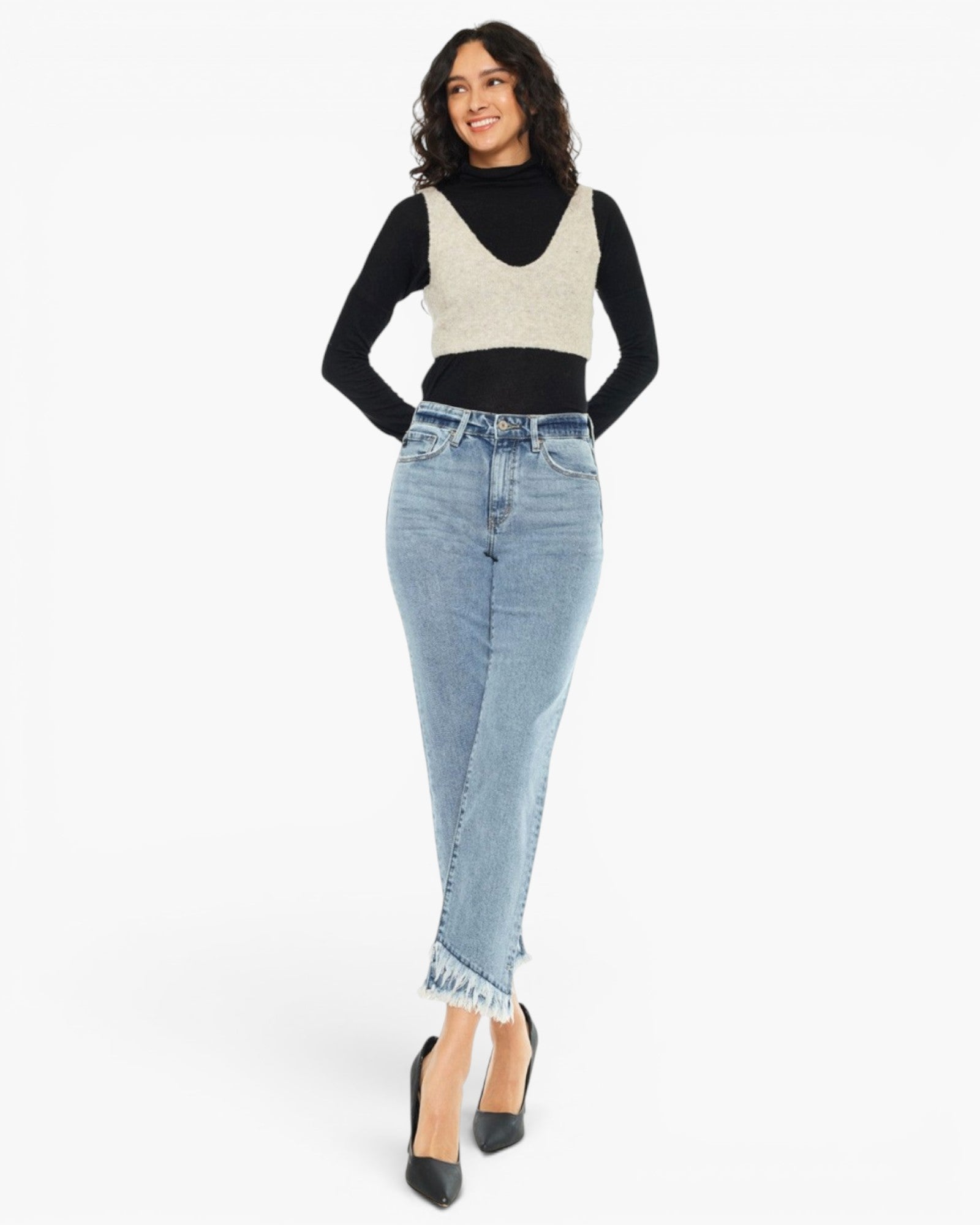 A woman modeling a light-wash blue denim midi skirt with frayed hems, paired with a black long-sleeve top and beige knit vest, against a plain white background.