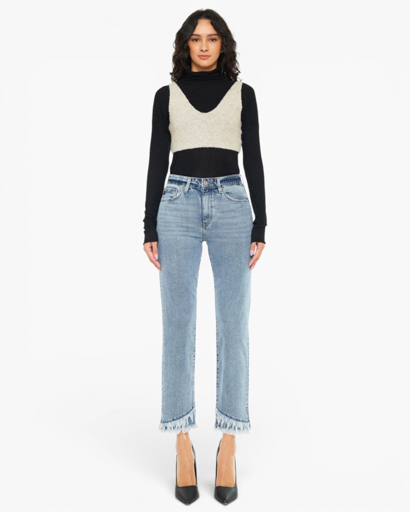 A model stands against a plain white studio background wearing a black long-sleeve turtleneck, a cream knit vest, light-wash blue cropped jeans with frayed hems, and black pointed heels.