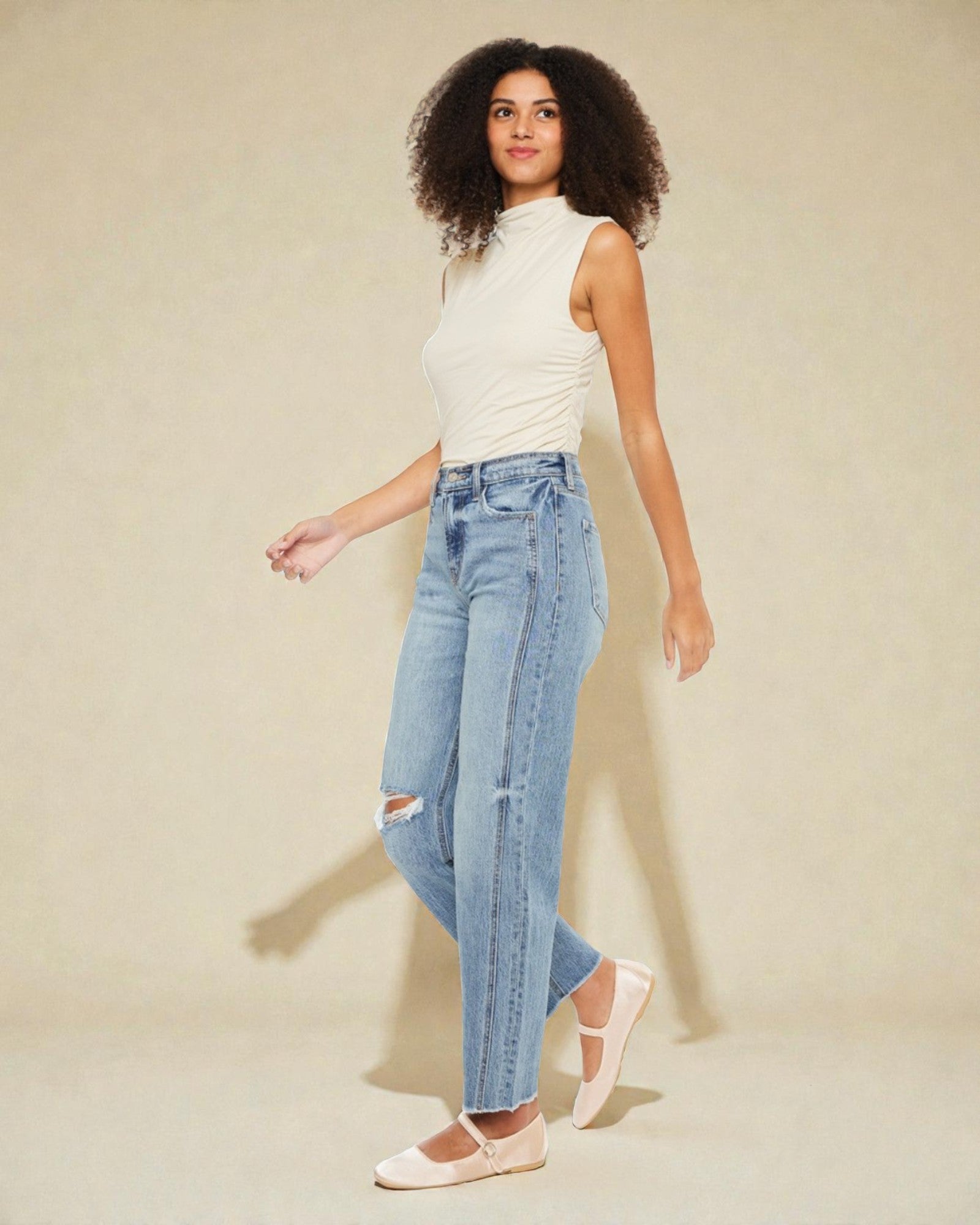 Woman wearing a white sleeveless top and light blue jeans against a beige background
