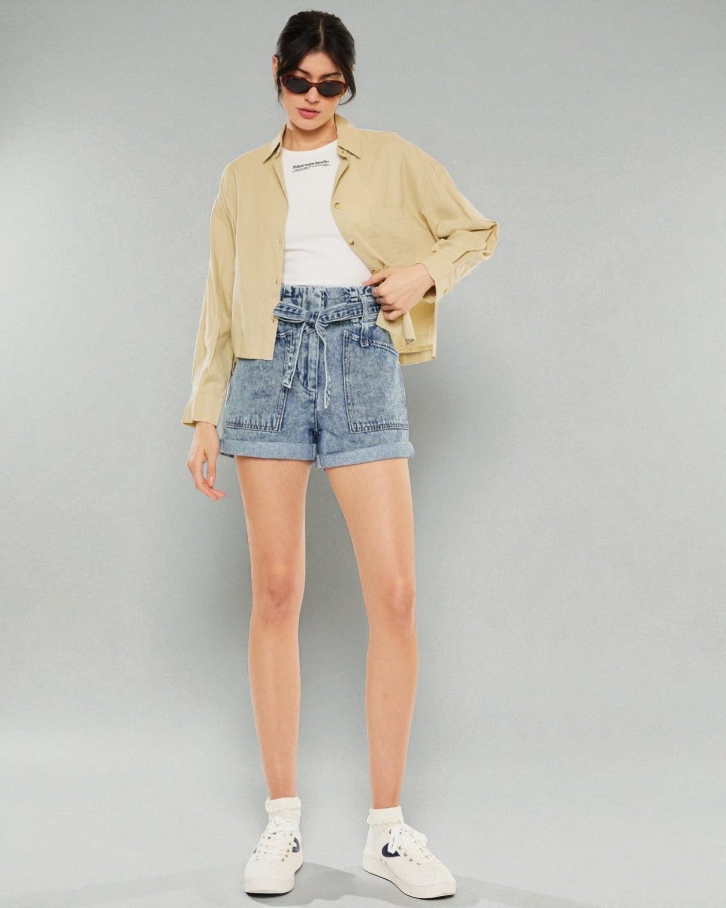 Woman wearing a beige shirt, white top, blue denim shorts, and white sneakers on a gray background