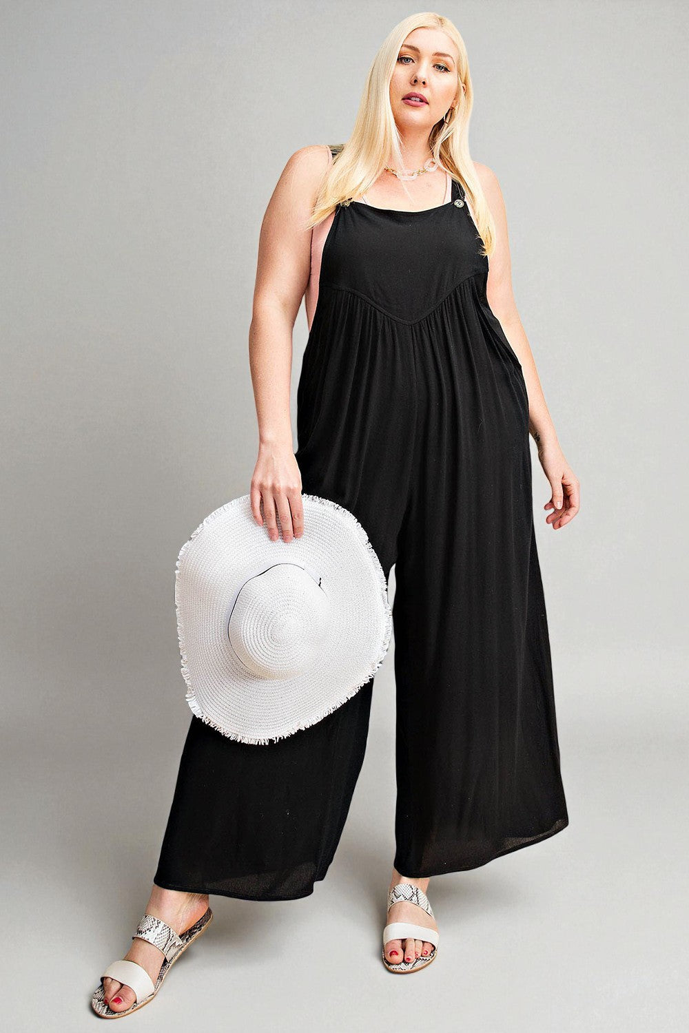 Woman wearing a black jumpsuit holding a white sun hat on a gray background