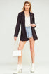 Woman wearing a black blazer, white top, denim shorts, and white boots on a light gray background