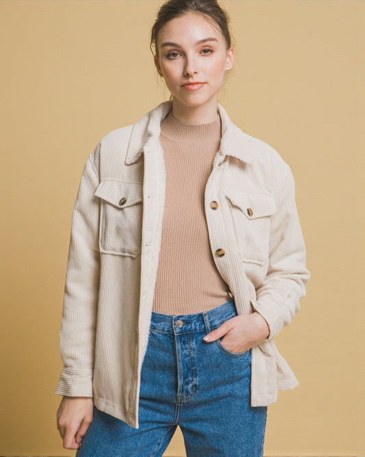 Woman wearing a beige jacket, pink sweater, and blue jeans against a beige background