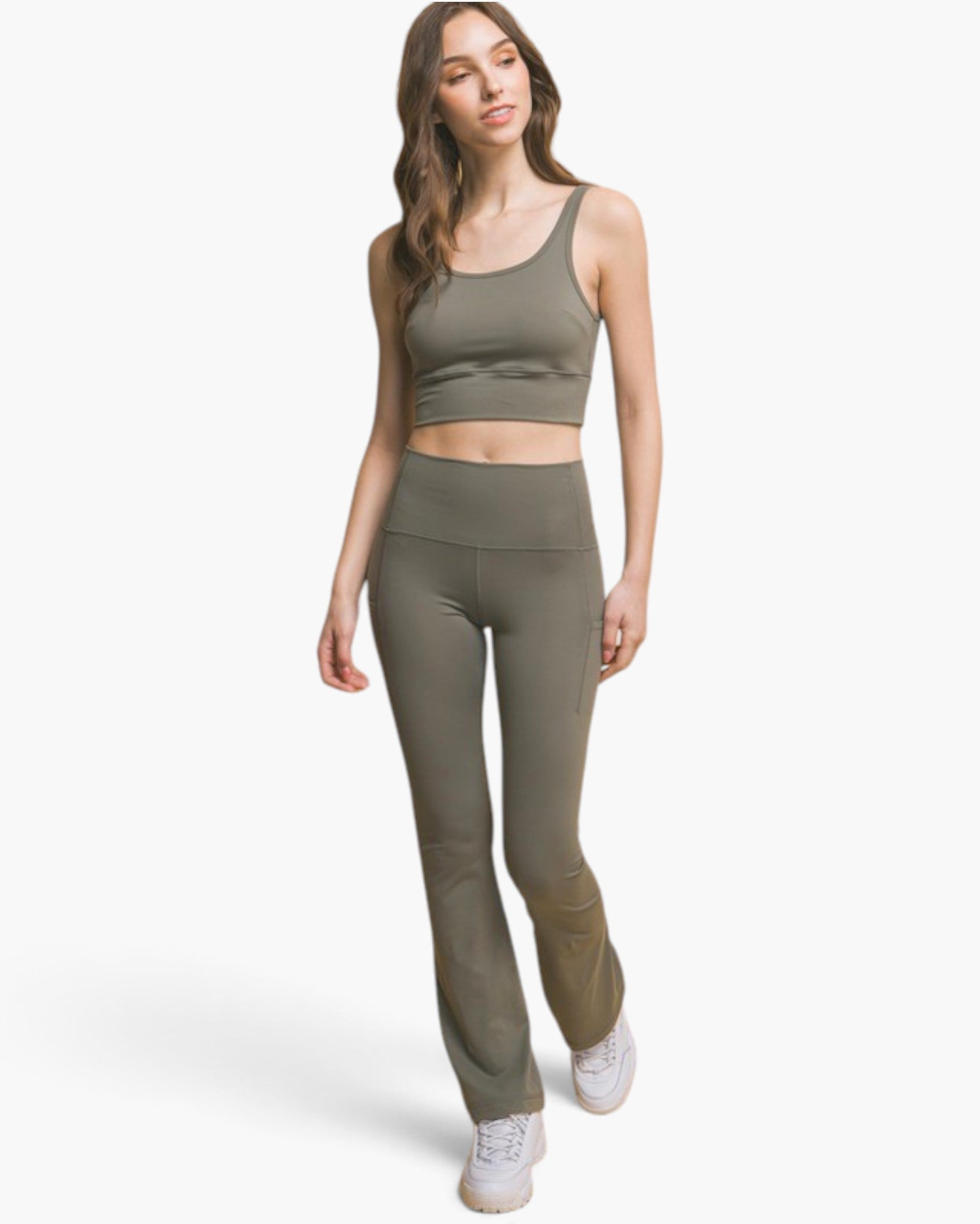 A woman modeling a matching olive green activewear set consisting of a cropped tank top and high-waisted flared leggings, paired with white sneakers, against a light gray studio background.