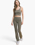 A woman modeling a matching olive green activewear set consisting of a cropped tank top and high-waisted flared leggings, paired with white sneakers, against a light gray studio background.