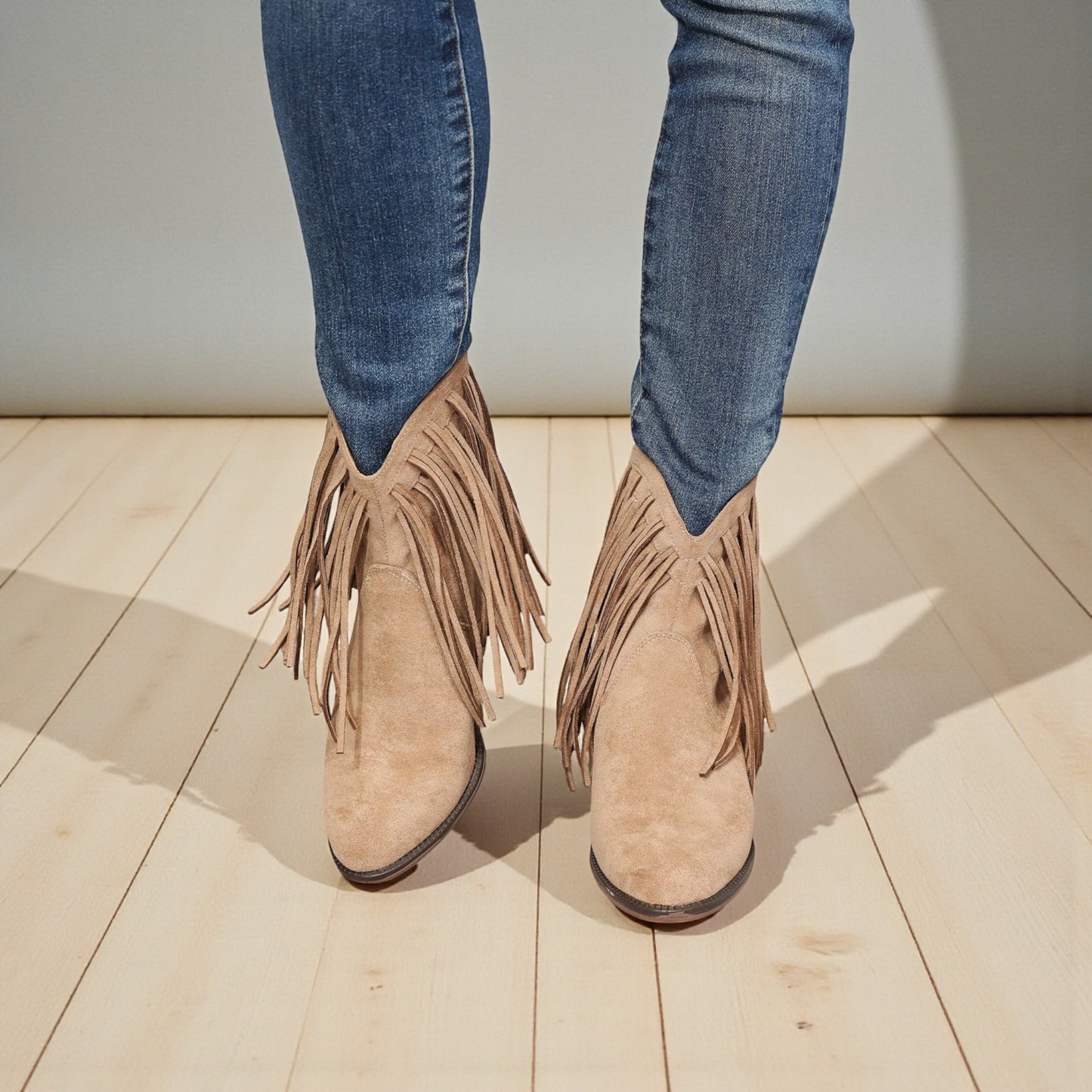 Beige fringed boots worn with blue jeans on a wooden floor.