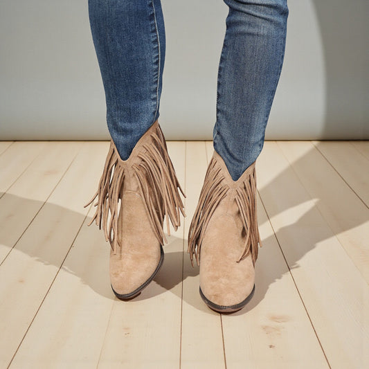 Beige fringed boots worn with blue jeans on a wooden floor.