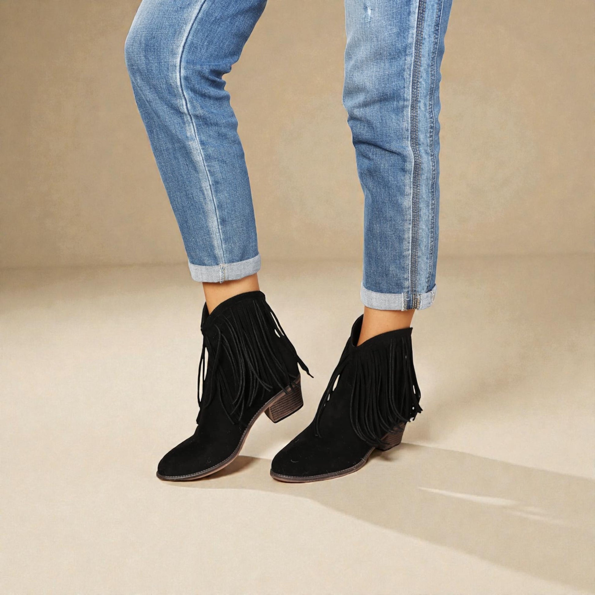 Black fringed ankle boots worn with rolled-up blue jeans on a beige background