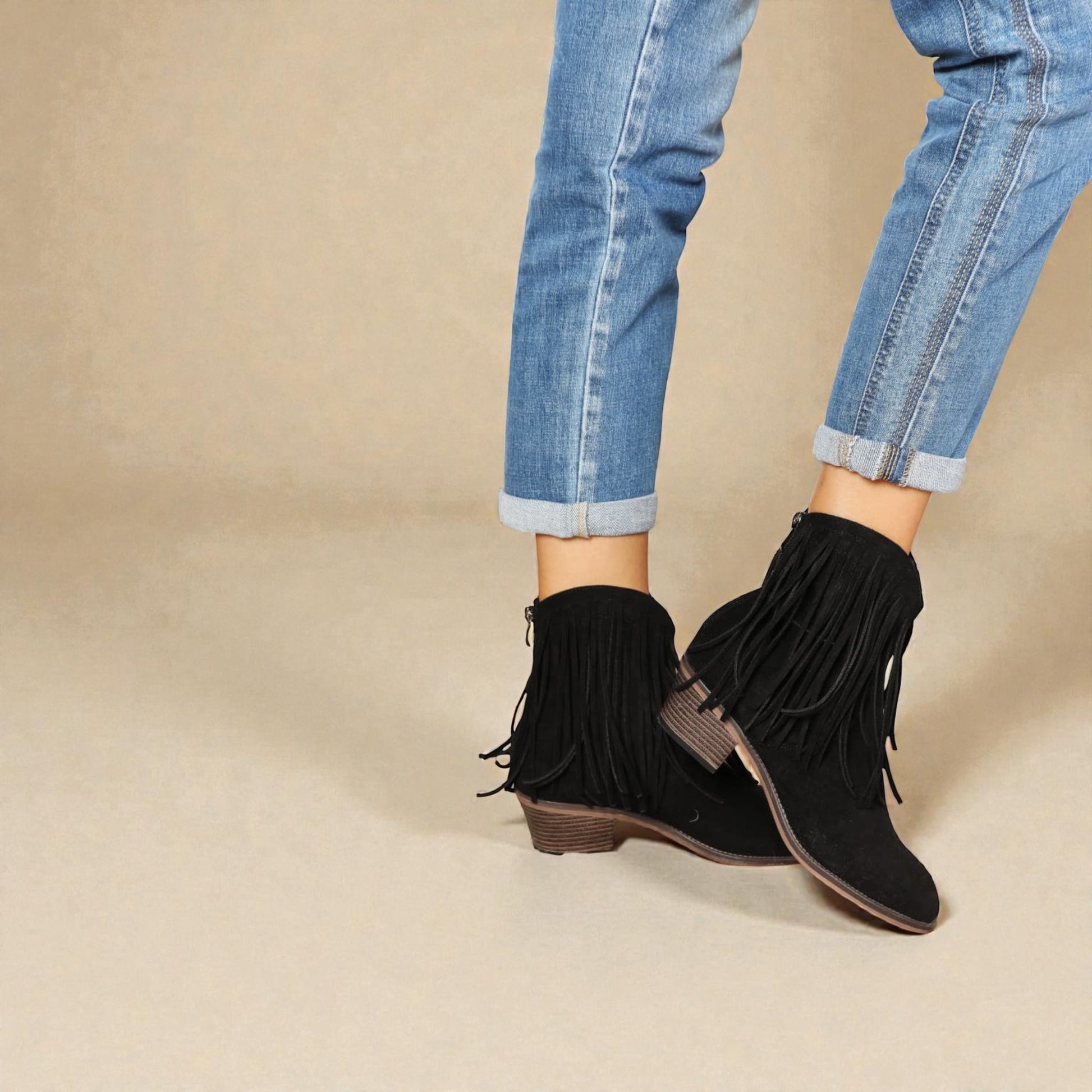 Black fringed boots worn with rolled-up blue jeans on a beige background