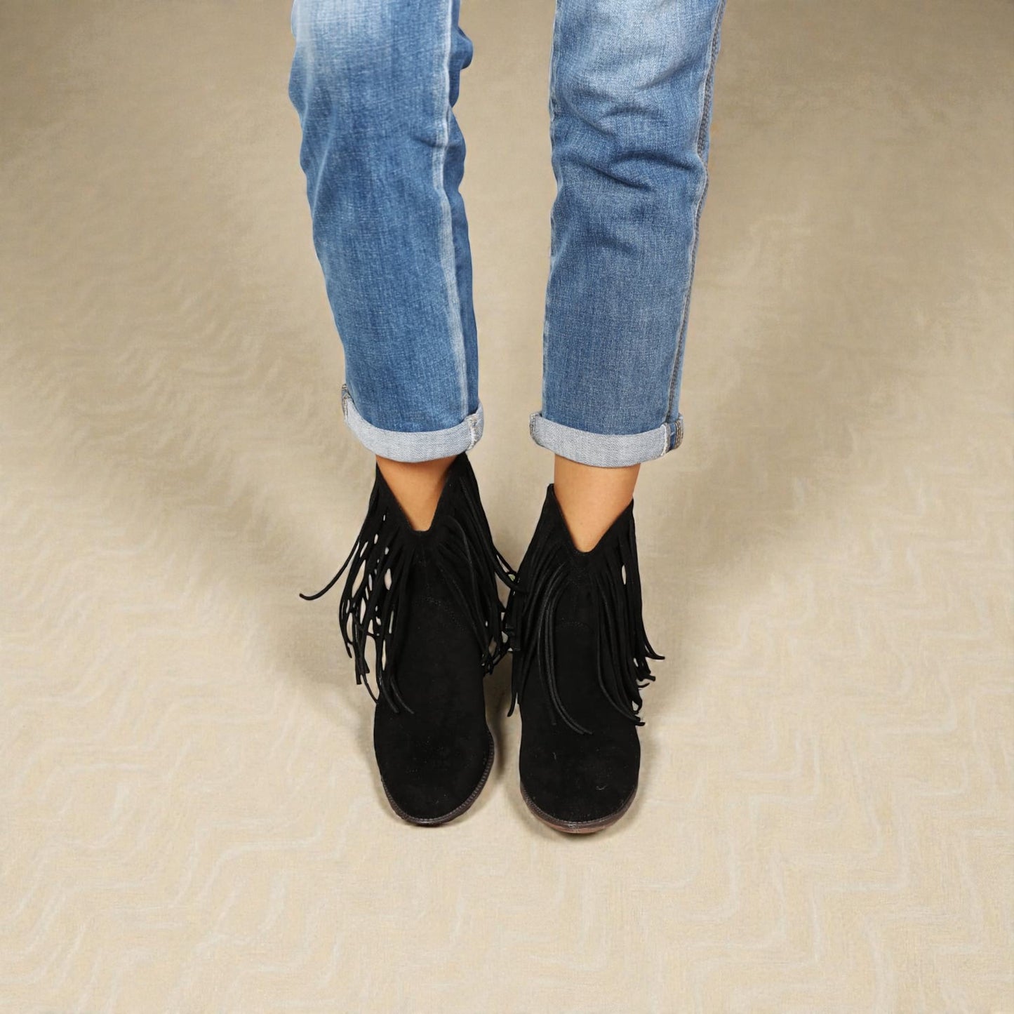 Black fringed boots worn with rolled-up blue jeans on a beige surface