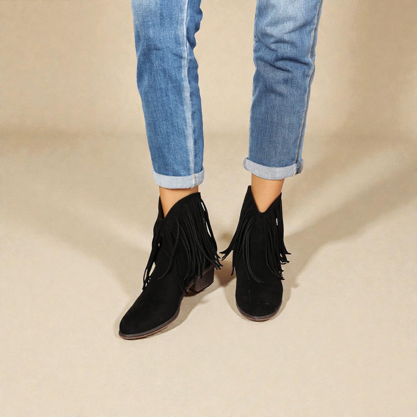 Black fringed ankle boots worn with rolled-up blue jeans on a beige background