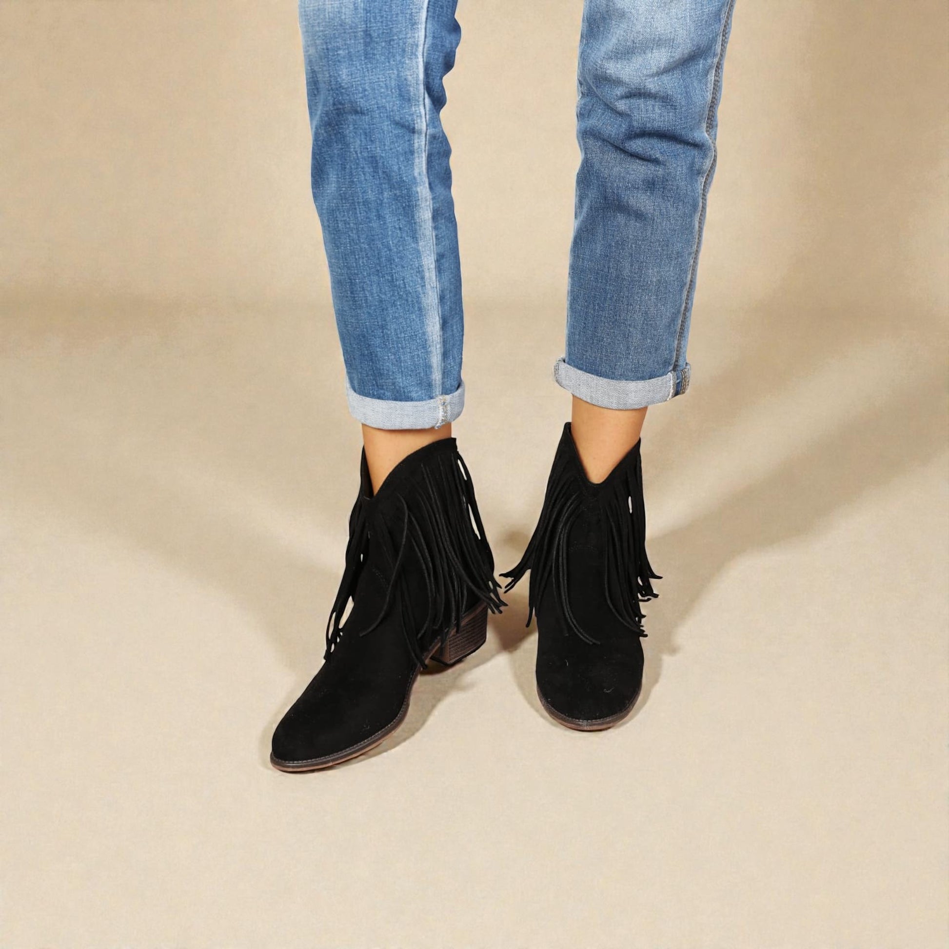 Black fringed ankle boots worn with rolled-up blue jeans on a beige background