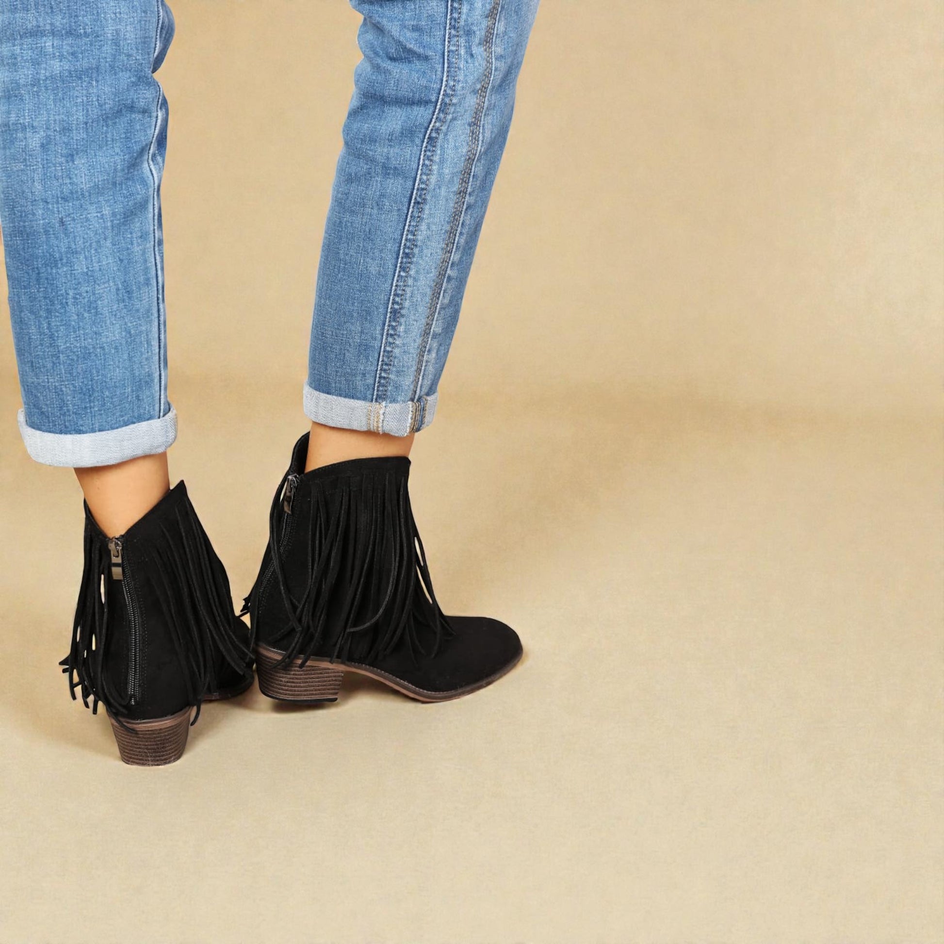 Black fringe ankle boots worn with rolled-up jeans on a beige background