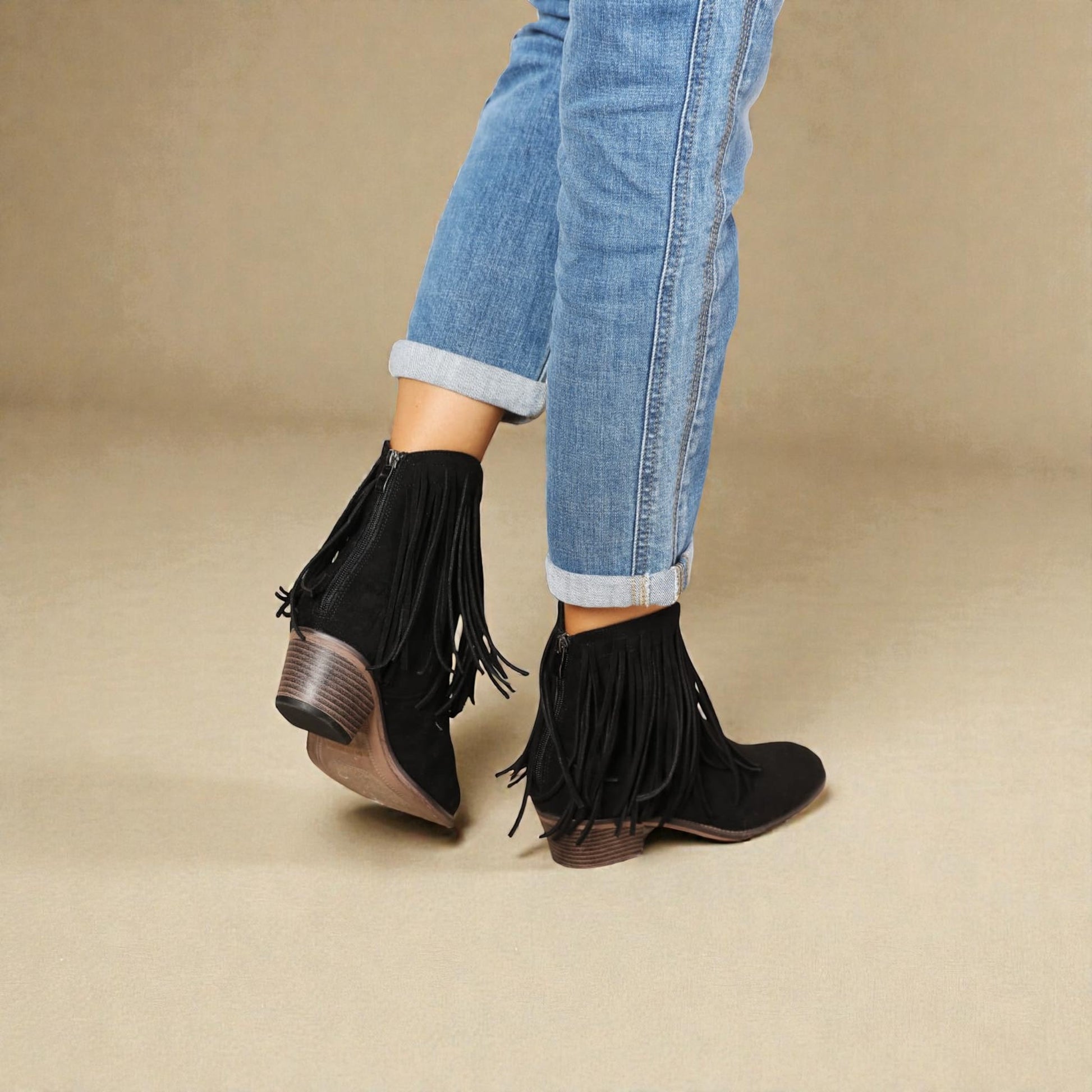 Black fringed ankle boots worn with rolled-up jeans on a beige background
