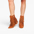 A pair of women's pointed-toe ankle boots in a warm cognac brown suede, featuring multiple tiers of fringe and a side zipper, shown on a model's feet against a white background.