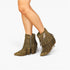 A pair of olive green suede fringe ankle boots with side zippers, shown on a model's feet against a plain white background.