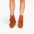 A pair of women's ankle boots in a warm brown suede-like material, featuring multiple tiers of fringe and a block heel, shown on a model's feet against a light gray background.