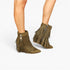 A pair of olive green fringe ankle boots with side zippers, shown on a model's feet against a plain white background.