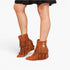 A pair of women's brown suede ankle boots with long fringe and a low block heel, shown on a model's feet against a light gray studio background.