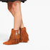A pair of women's suede ankle boots in a warm rust-brown color, featuring long fringe detailing and a silver zipper pull, shown on a model's feet against a plain white background.