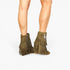 A pair of olive green suede fringe ankle boots with side zippers, shown on a model's feet against a plain white background.