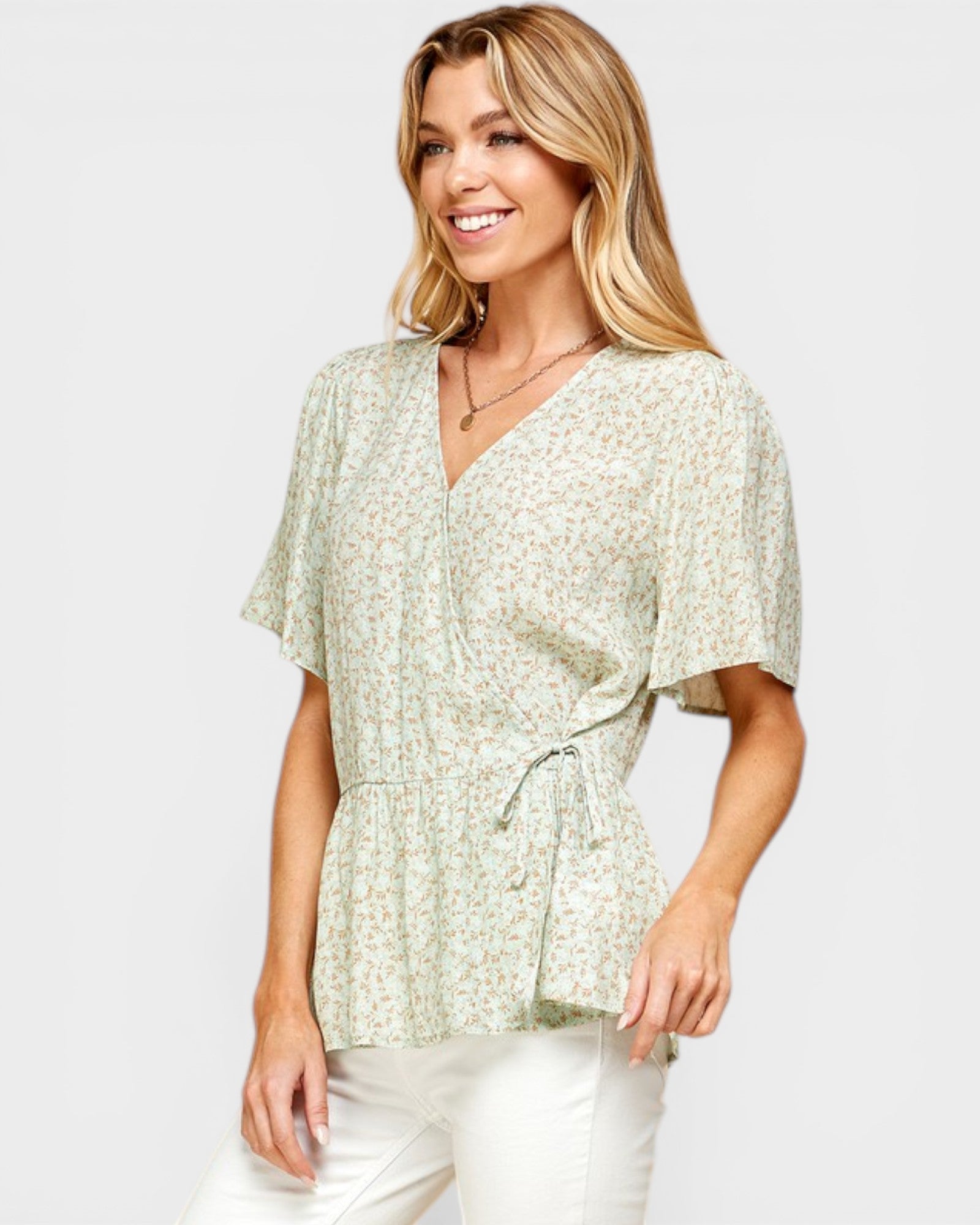Woman wearing a light green patterned blouse with white pants on a plain background