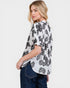 Woman wearing a white blouse with black floral pattern and dark jeans on a plain background