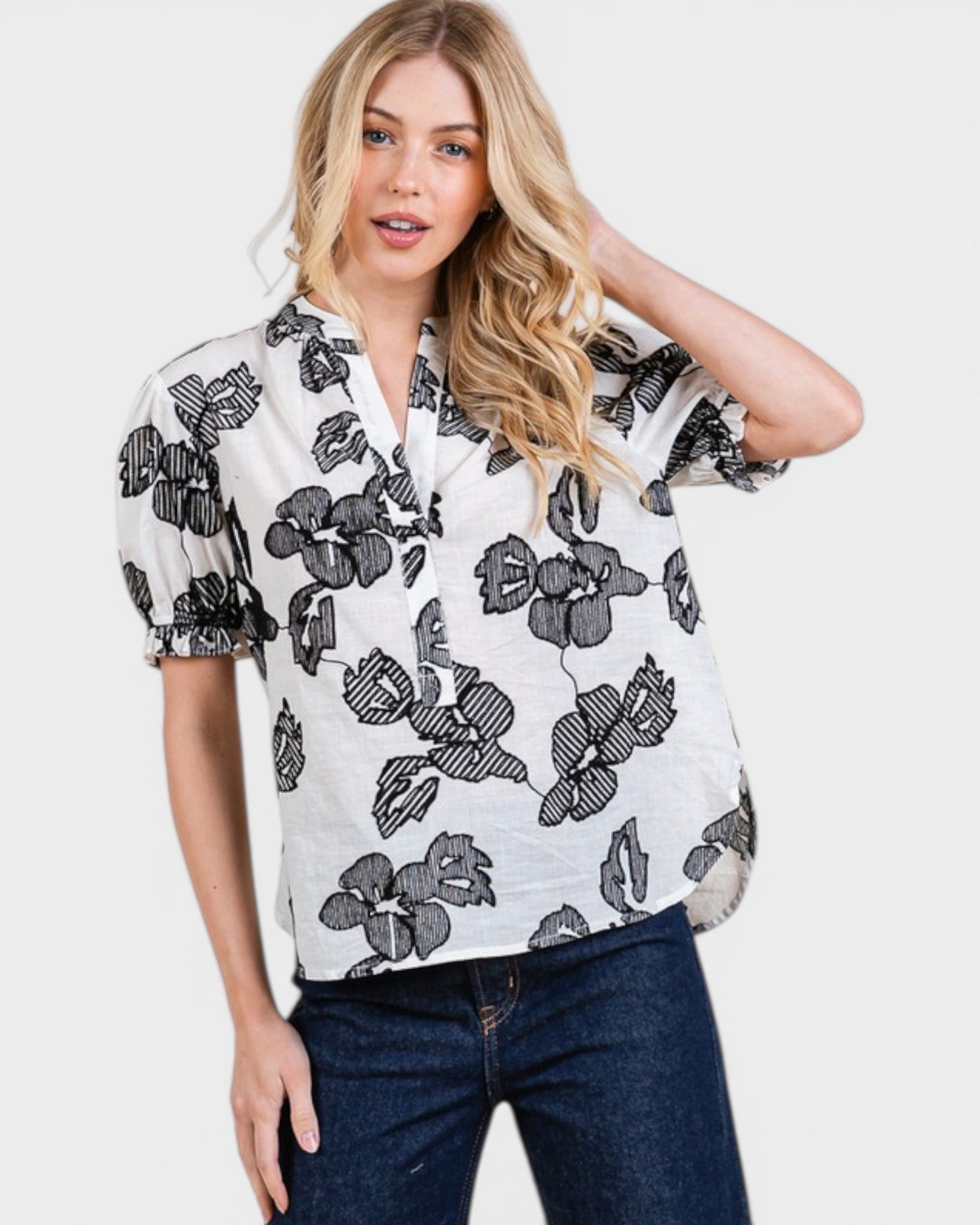 Woman wearing a white blouse with black floral pattern and blue jeans on a plain background