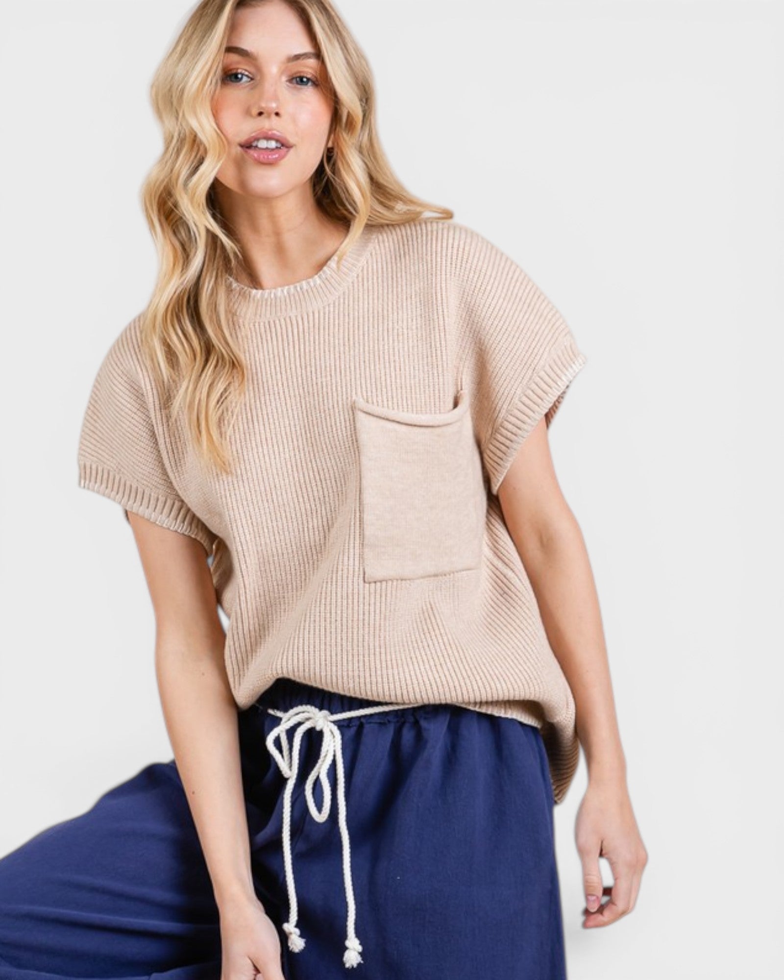 Woman wearing a beige knit top with a pocket and navy pants on a white background