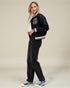 Woman wearing a black varsity jacket with white lettering, black pants, and blue sneakers on a beige background