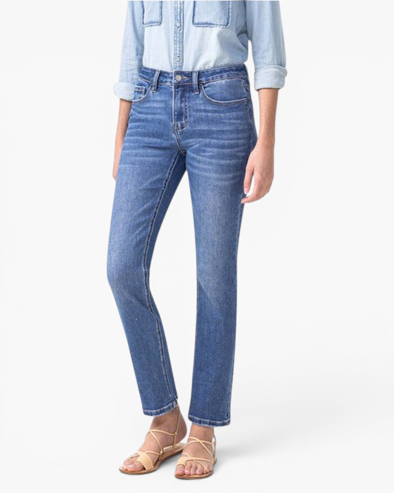 A model wearing a pair of medium-wash blue denim jeans with a straight-leg fit, styled with a light blue denim shirt and beige strappy sandals against a plain white background.