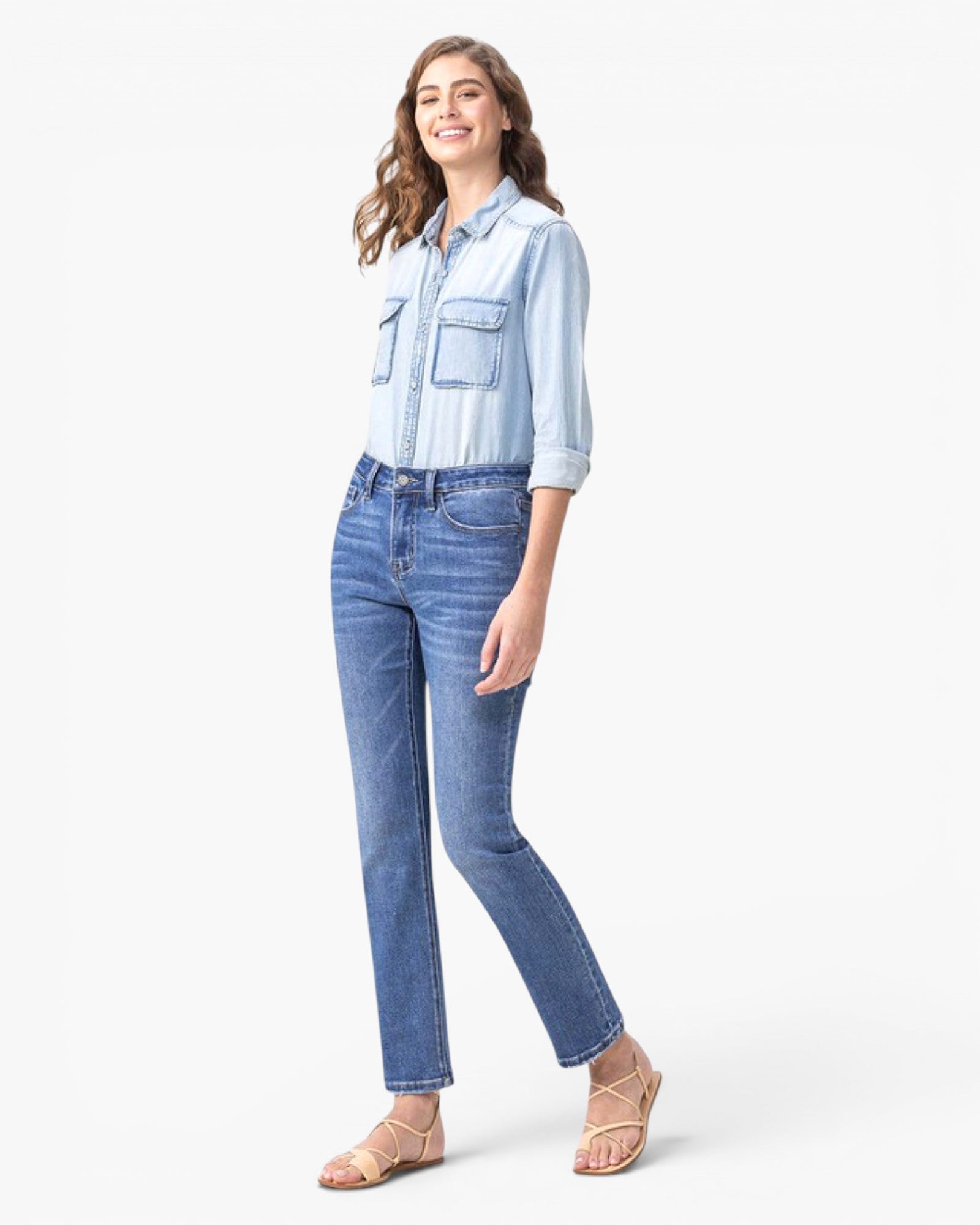 A woman modeling a light blue denim button-up shirt and medium-wash blue jeans with beige strappy flat sandals, standing against a plain white studio background.
