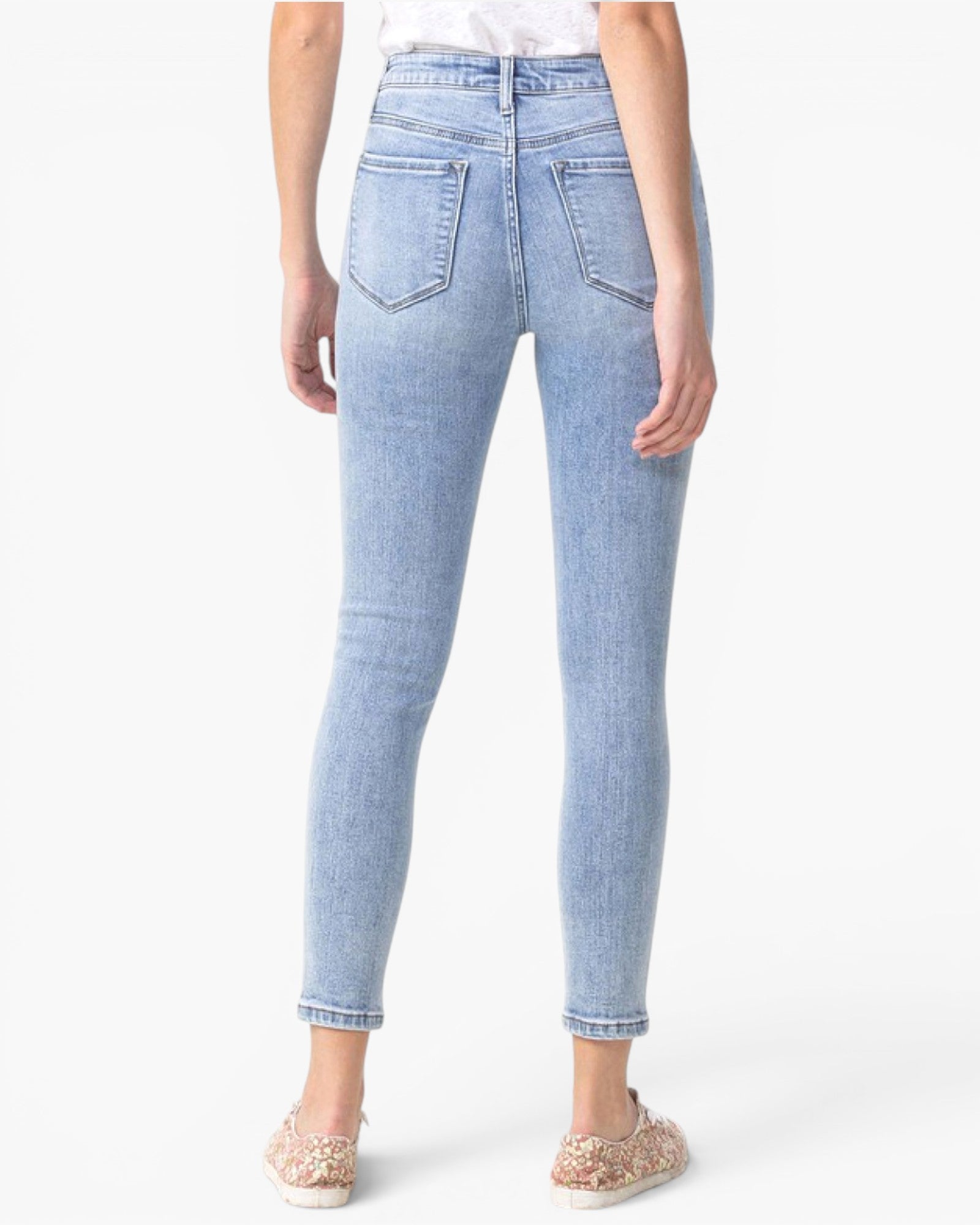A person shown from the back wearing light-wash blue skinny jeans with rolled cuffs and flat glittery slip-on shoes, against a plain white studio background.