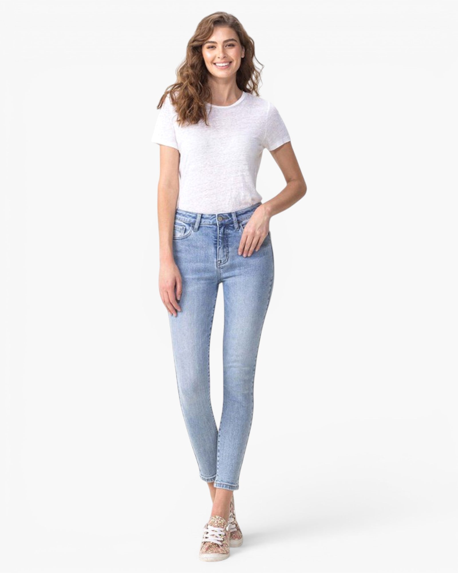 A woman modeling light-wash blue skinny jeans with a white short-sleeve t-shirt and white sneakers, standing against a plain white studio background.