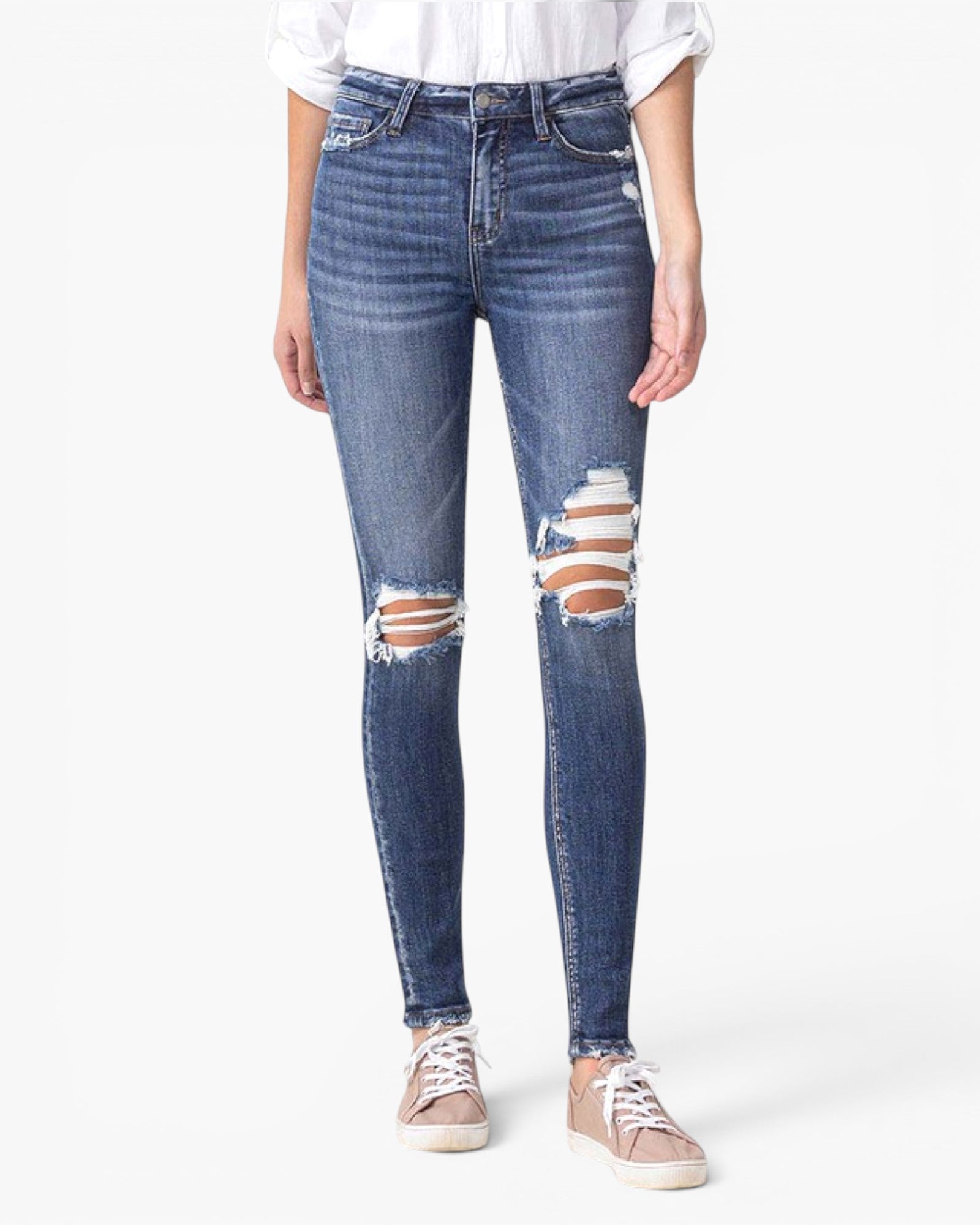 A person wearing blue distressed skinny jeans with ripped knees, paired with a white top and beige sneakers, shown against a plain light gray studio background.