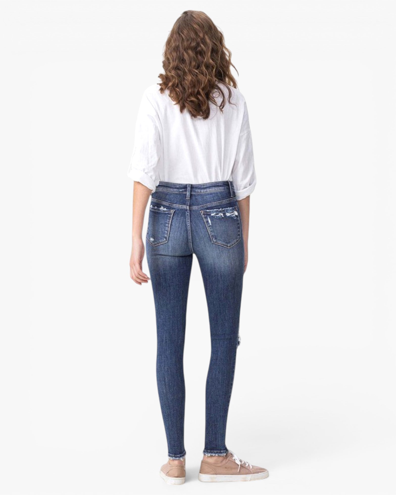 A woman shown from the back wearing blue skinny jeans with a white shirt and beige sneakers, photographed against a plain white studio background.